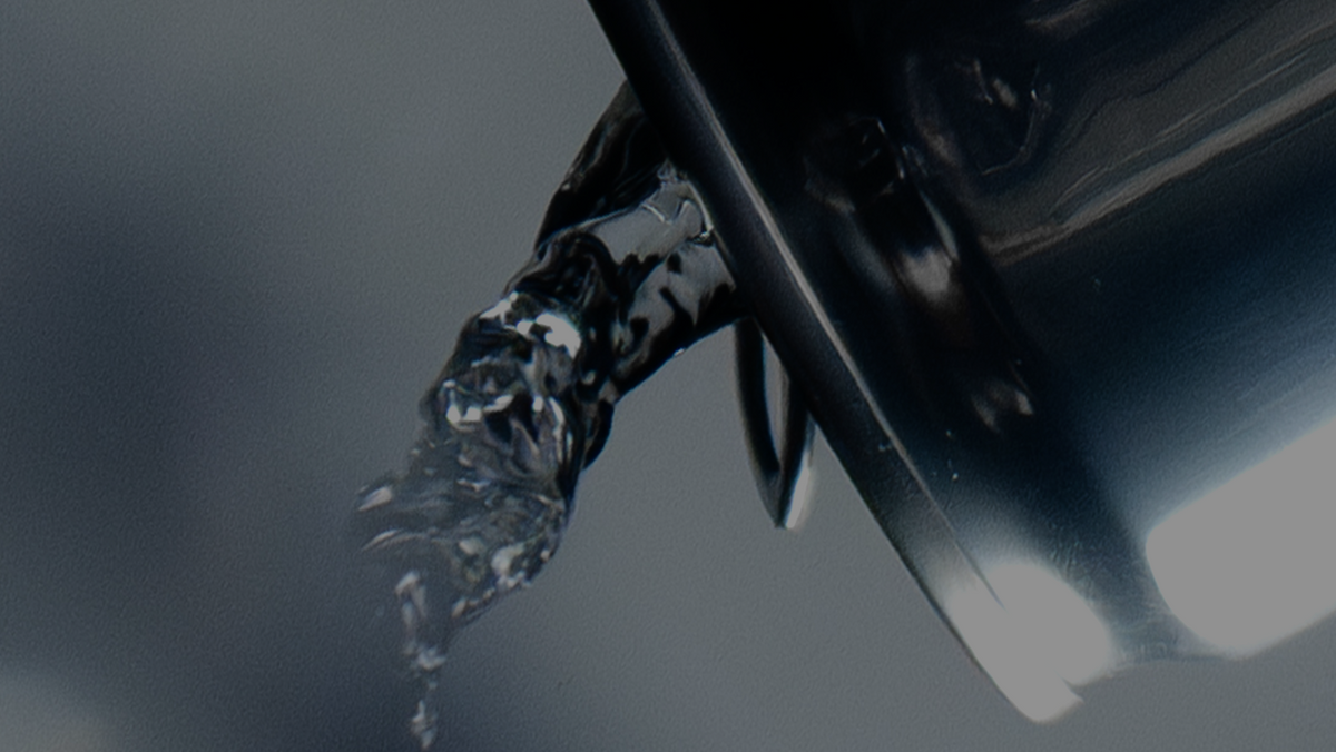 A close-up, dramatic shot of a clear liquid splash bursting from the open top of a glossy black container against a dark gray background.